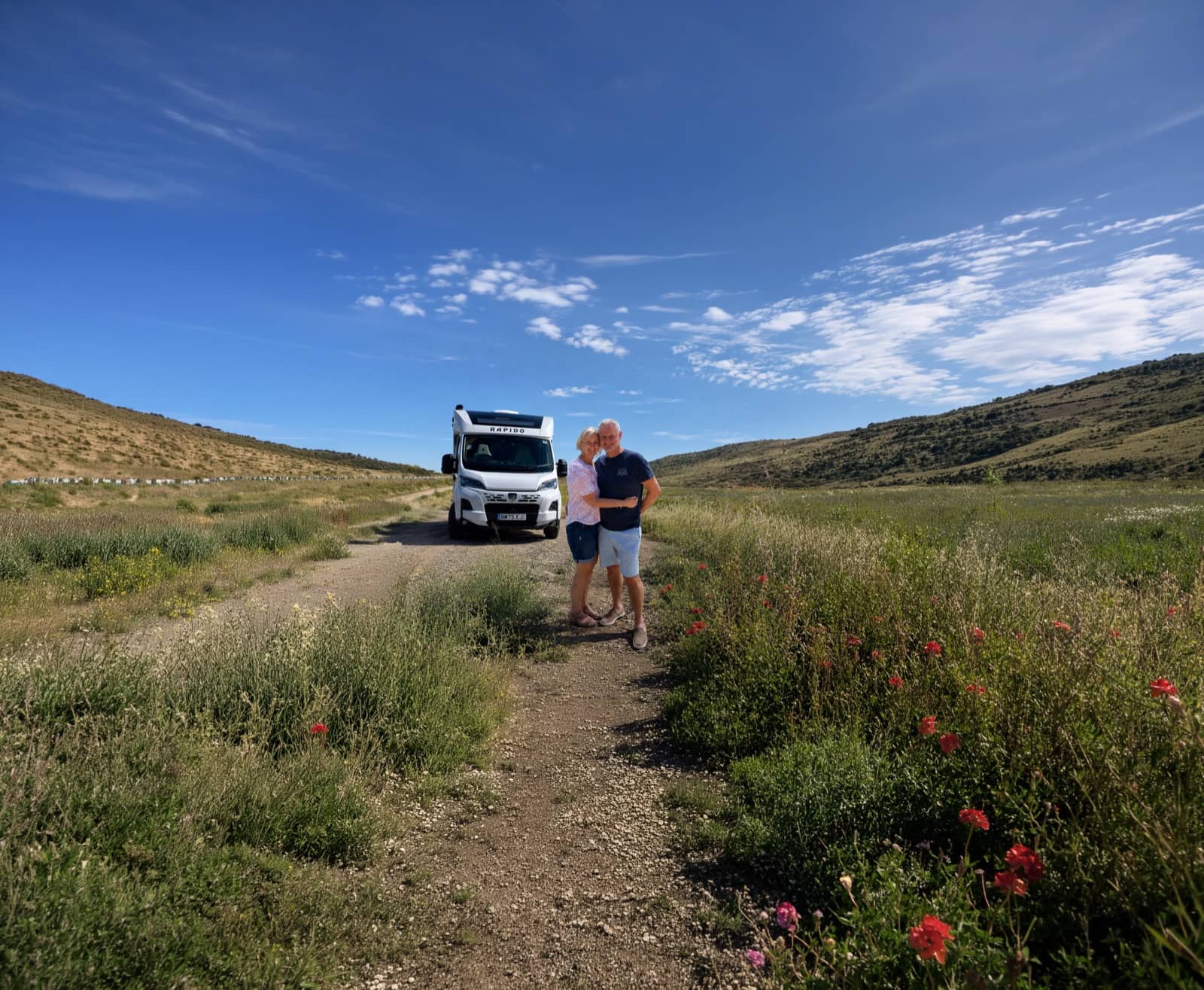 Una pareja de pie junto a su autocaravana en un camino que atraviesa colinas verdes